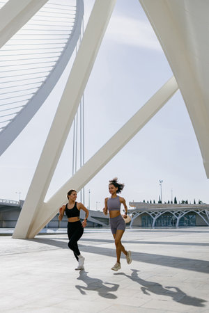 Two active women athlete running side by side along an outdoor track on modern buildings background