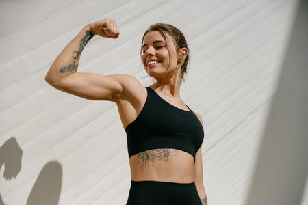 Smiling woman athlete raising arm and showing biceps while standing on modern buildings background