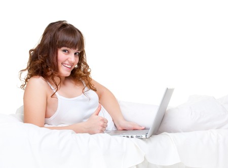 smiley woman lying in white bed with laptop and showing thumbs upの写真素材