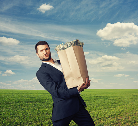 young businessman holding heavy paper bag with money over blue sky and green fieldの写真素材