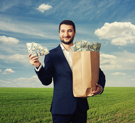 smiley handsome businessman holding paper bag with money and showing dollars.  photo over blue sky and green fieldの写真素材