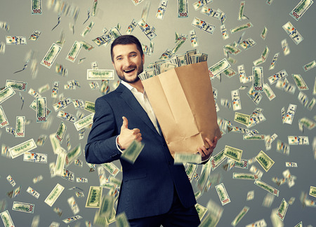 excited happy businessman standing under dollar's rain and showing thumbs upの写真素材