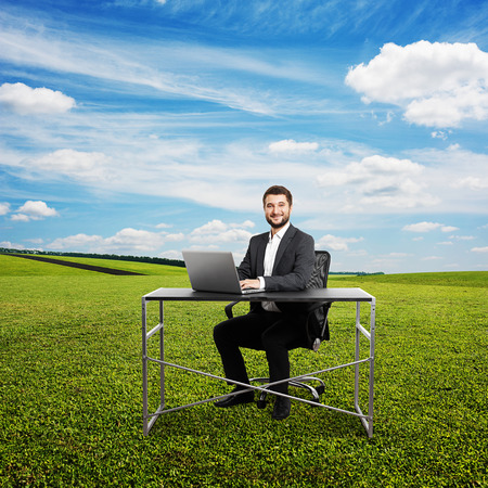 successful smiley businessman sitting at the table, typing at laptop and looking at camera. photo at outdoorの写真素材