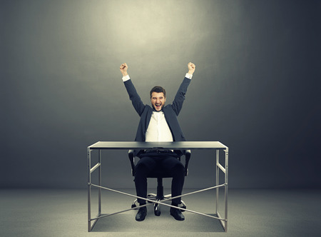 excited businessman sitting at the table, raising hands up up and laughing. photo in the dark roomの写真素材