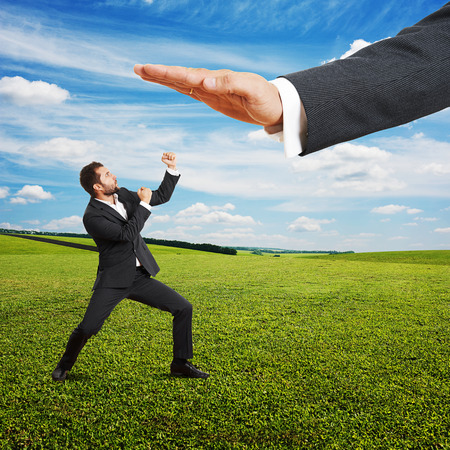 concept photo of conflict between subordinate and boss. angry young businessman showing fist and looking up at big palm of his boss. photo at outdoorの写真素材