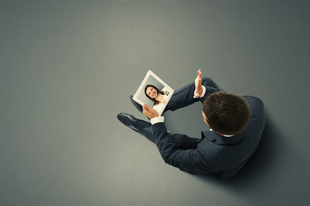 businessman sitting on the floor, holding tablet pc and waving his hand to young smiley woman. photo in the dark roomの写真素材