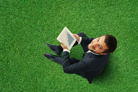 top view of smiley businessman sitting on the green grass and holding touch pad with growing graphの写真素材