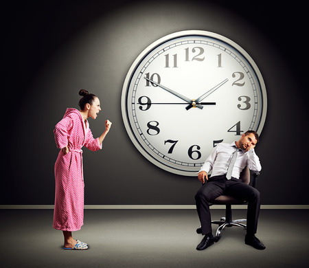 angry wife screaming at lazy husband. photo in dark room with big clock on the wallの写真素材