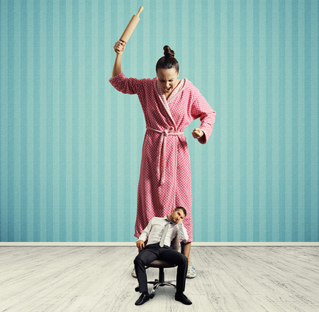 dissatisfied woman with rolling pin screaming at small lazy husband in room with blue wallの写真素材