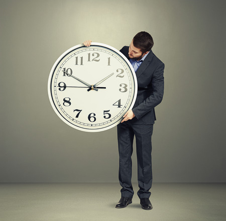 businessman holding big white clock and looking at clock dial over grey backgroundの写真素材