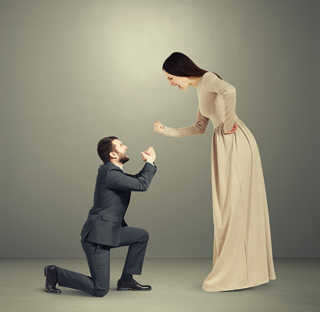 full length portrait of emotional couple over grey background. woman screaming and showing fist, man standing on knee and apologizingの写真素材