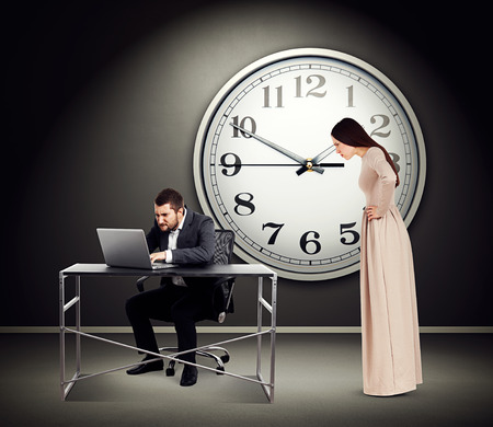 young beautiful woman staring at man with laptop. photo in dark room with big white clock on the wallの写真素材