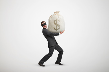 laughing man in formal wear and black mask on the eyes holding big bag with money and looking at camera against light grey backgroundの写真素材