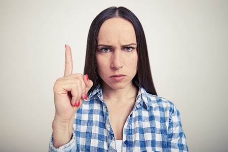 portrait of serious young woman showing attention sign and looking at camera over light grey backgroundの写真素材