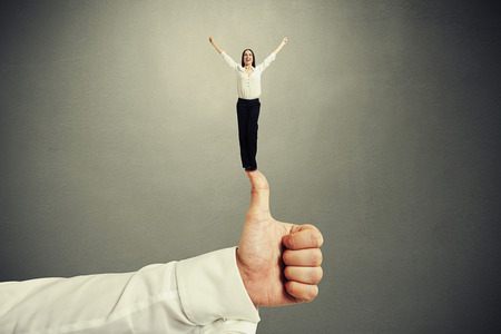happy small woman standing on big thumbs up and raising her hands up over dark grey backgroundの写真素材