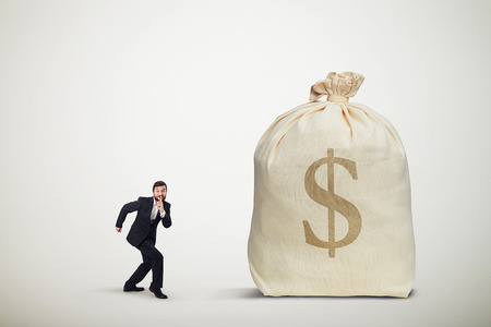 businessman in formal wear showing silent sign and sneaking to the big bag of money over light grey backgroundの写真素材