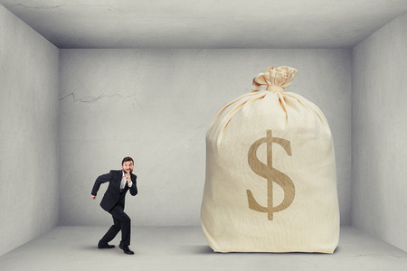 businessman in formal wear showing silent sign and walking on tiptoe to the big bag of money in grey roomの写真素材