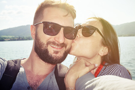 happy kissing couple taking selfie against beautiful view with lake and hills