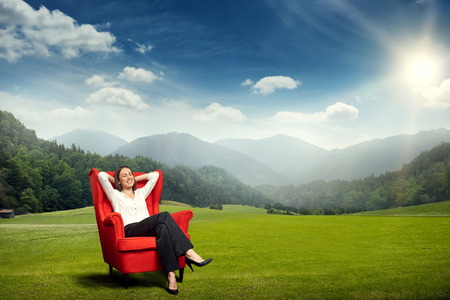 smiley young woman resting in red chair on the green meadow over beautiful landscape with hills, forest and skyの写真素材