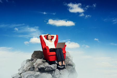 smiley businesswoman with laptop on the red chair resting on top of rock over blue skyの写真素材