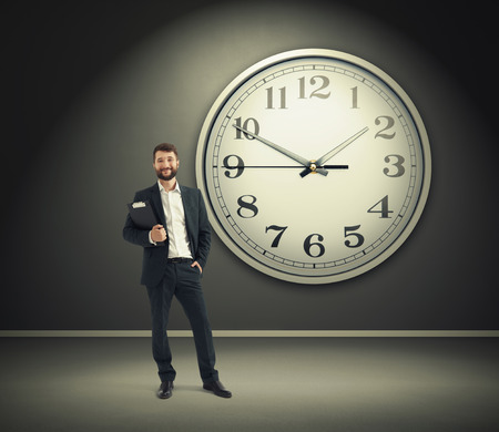 successful businessman holding black folder and looking at camera over big white clock on grey wallの写真素材