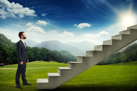 smiley businessman standing on the green meadow and looking up to the stairsの写真素材