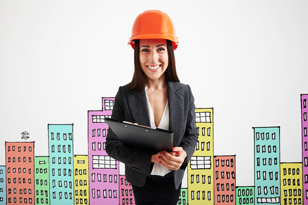 smiley businesswoman in orange helmet holding black folder against variegated drawing cityscapeの写真素材