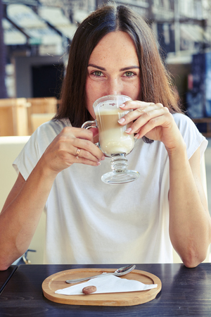 cheerful woman in white t-shirt sitting in cafe, drinking cappuccino and looking at cameraの写真素材