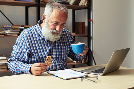 smiley senior businessman holding cup of coffee and cookies and looking at notepadの写真素材