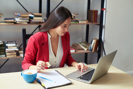 young woman working with laptop and eating cookies in office