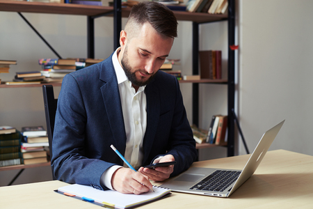 young businessman holding his smartphone and writing on notepad in officeの写真素材