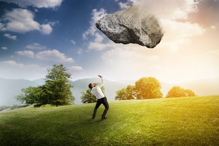 scared businessman under big flying stone over bright landscapeの写真素材