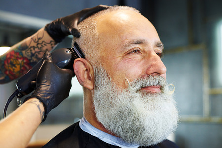 sideview portrait of handsome bearded man in professional barbershop. barber shaving beard with electric razorの写真素材