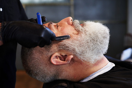 portrait of senior man in barbershop. barber shaving man with straight razorの写真素材