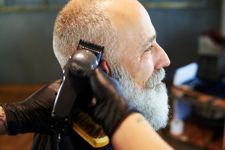 sideview portrait of senior bearded man in professional barbershop. barber shaving beard with electric razorの写真素材
