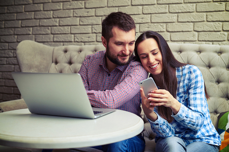 smiley young couple sitting on sofa and using gadgetsの写真素材