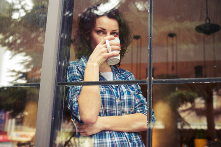 beautiful young woman standing near window, drinking the coffee and looking into the distance