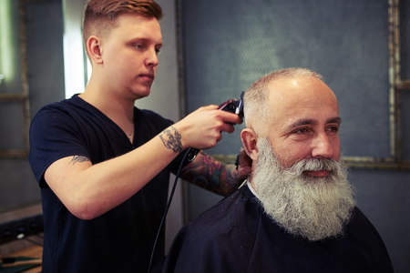 Concentrated skillful barber making haircut to handsome attractive senior man with beard and working with electric razorの写真素材