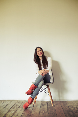 Smiling woman is sitting on a chair and posing, background wooden floor and white wallの写真素材