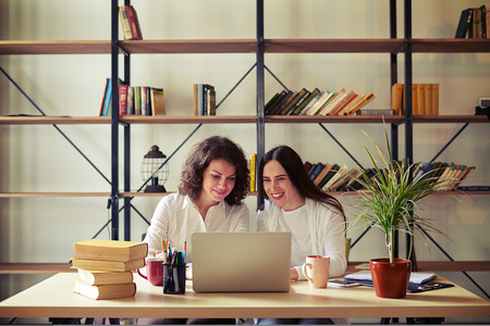 Two women sitting at the table and working at a laptop, bookshelves on a background