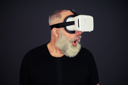 Beard senior man surprised using virtual reality glasses, on black backgroundの写真素材
