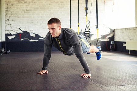 A young sportsman in training suite is standing in plank on the suspension trainer