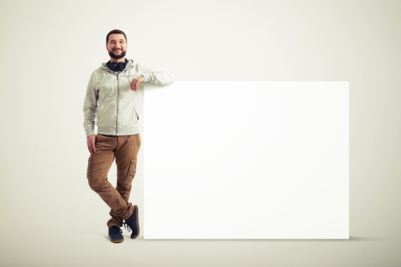Smiling young Caucasian bearded man in casual clothes is standing with his elbow resting on a huge white poster isolated on white backgroundの写真素材