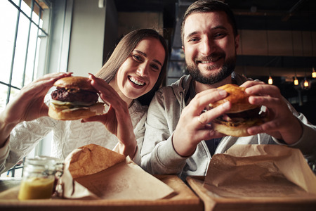A happy smiling woman and a handsome bearded man are enjoying their delicious and tasty burgers in the burger-barの写真素材