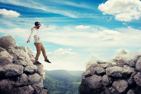 Man in virtual reality glasses is stepping carefully to walk on air between two high rocks on sunny valley