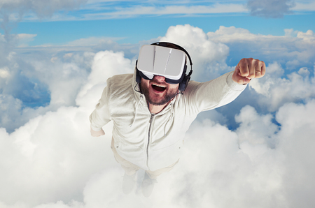 A bearded young man in white clothes is wearing virtual reality glasses and flying up in blue sky among white cloudsの写真素材