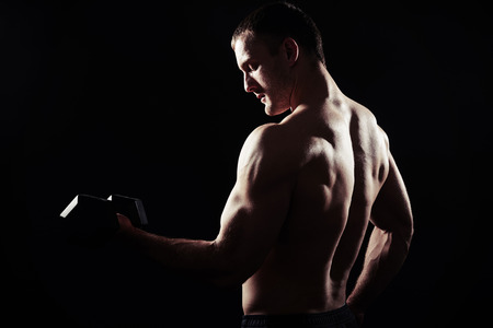 Bare-chested sexy muscular man is standing over black background with dumbbell in his hand, view from the backの写真素材