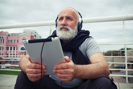Stylish aged man in headphones is sitting on a sidewalk on a cloudy day with tablet computer in his hands and musingの写真素材