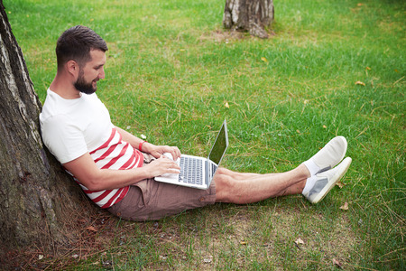 Young bearded man is sitting on green grass under the tree on the garden and working in his laptopの写真素材