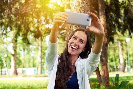 Beautiful young lady is smiling trying to make a selfie against a background of sunlit parkの写真素材